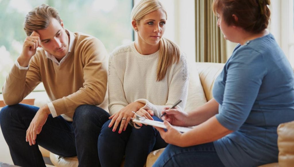 Couple engaged in marriage counseling session with therapist, discussing relationship challenges and seeking guidance for infidelity issues, in a comfortable therapy setting.