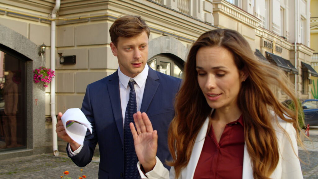 Man in a suit holding a piece of paper, gesturing towards a woman in a white blazer, both appearing to have a serious conversation in an outdoor setting, reflecting relationship dynamics during political discussions.