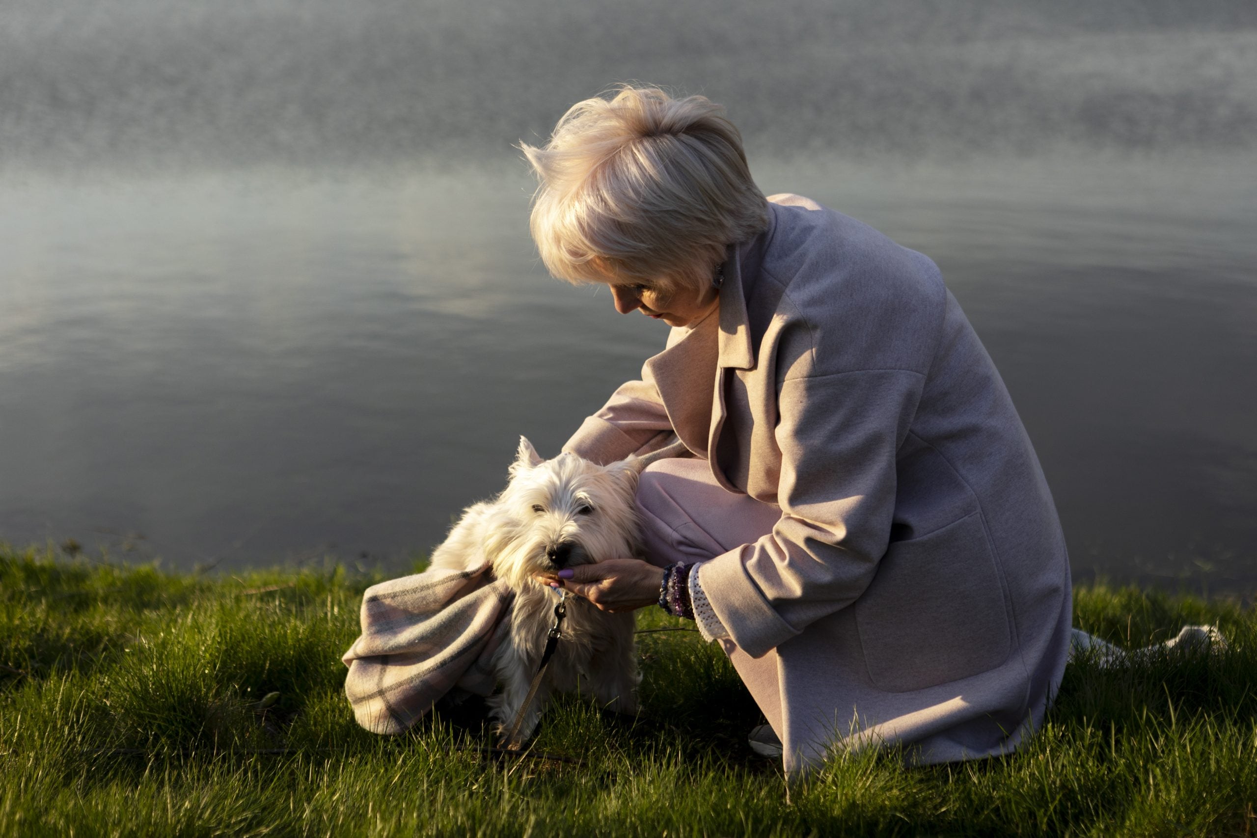 Woman comforting a small white dog by a serene waterfront, symbolizing the bond between pet parents and their pets in the context of grief and healing after pet loss.