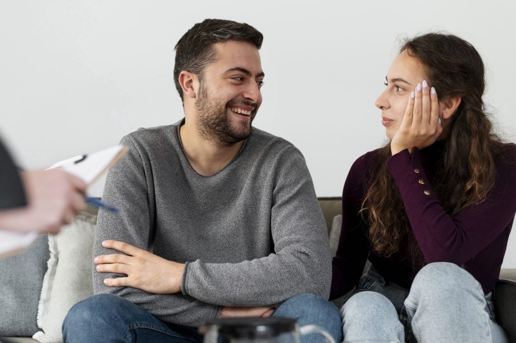 Couple engaged in counseling session, discussing relationship issues, therapist's hand with notepad visible, supportive atmosphere.