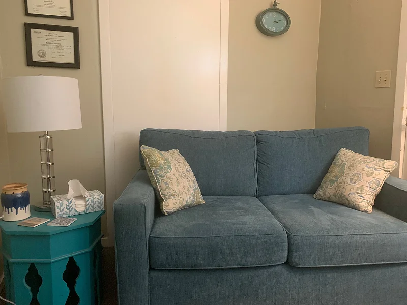 Cozy therapy office setting featuring a blue couch with decorative pillows, a lamp, a teal side table with tissues, and framed certificates on the wall, reflecting a welcoming atmosphere for counseling sessions.