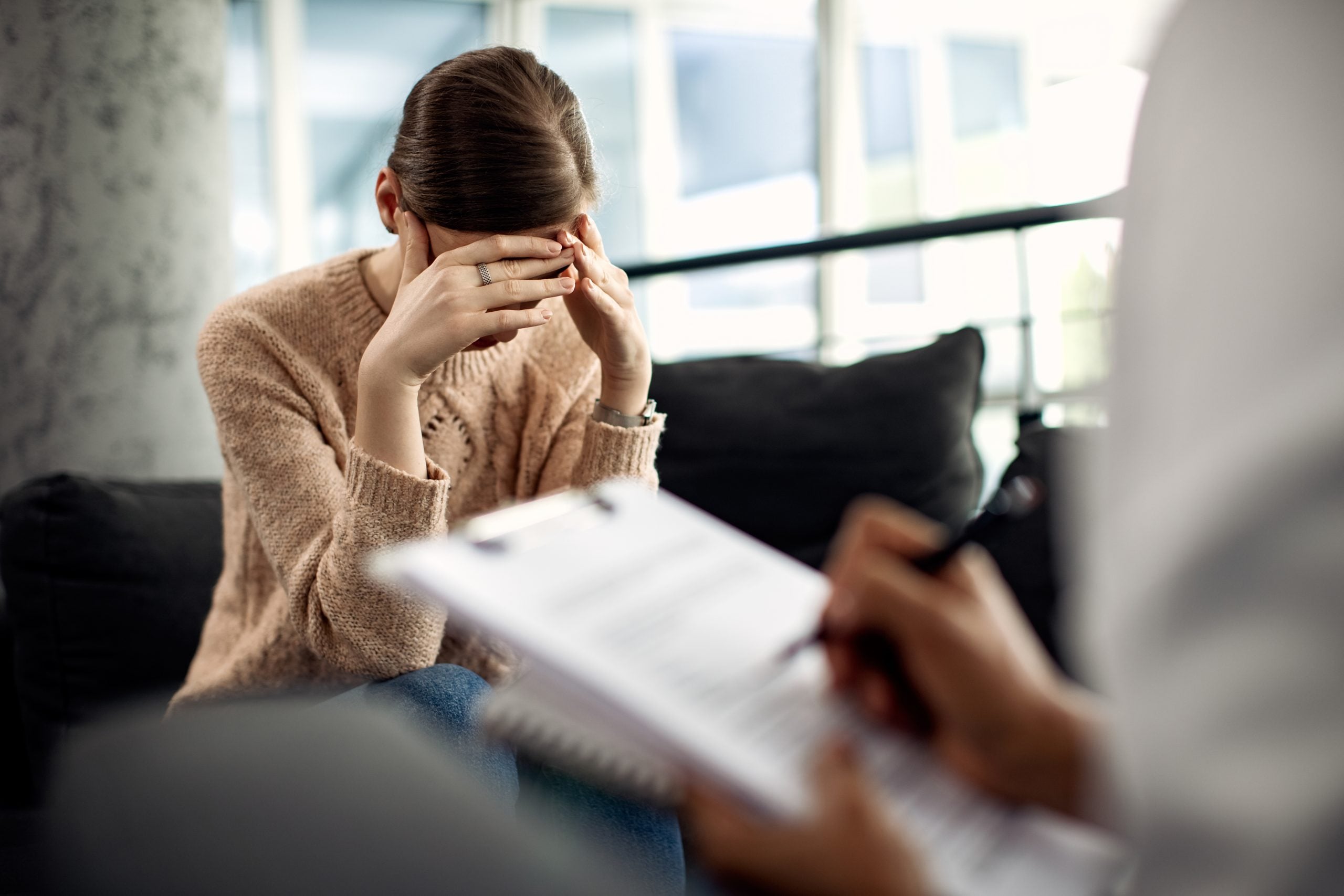 Woman sitting on a couch with hands on her face, experiencing emotional distress during psychotherapy session, while therapist takes notes.